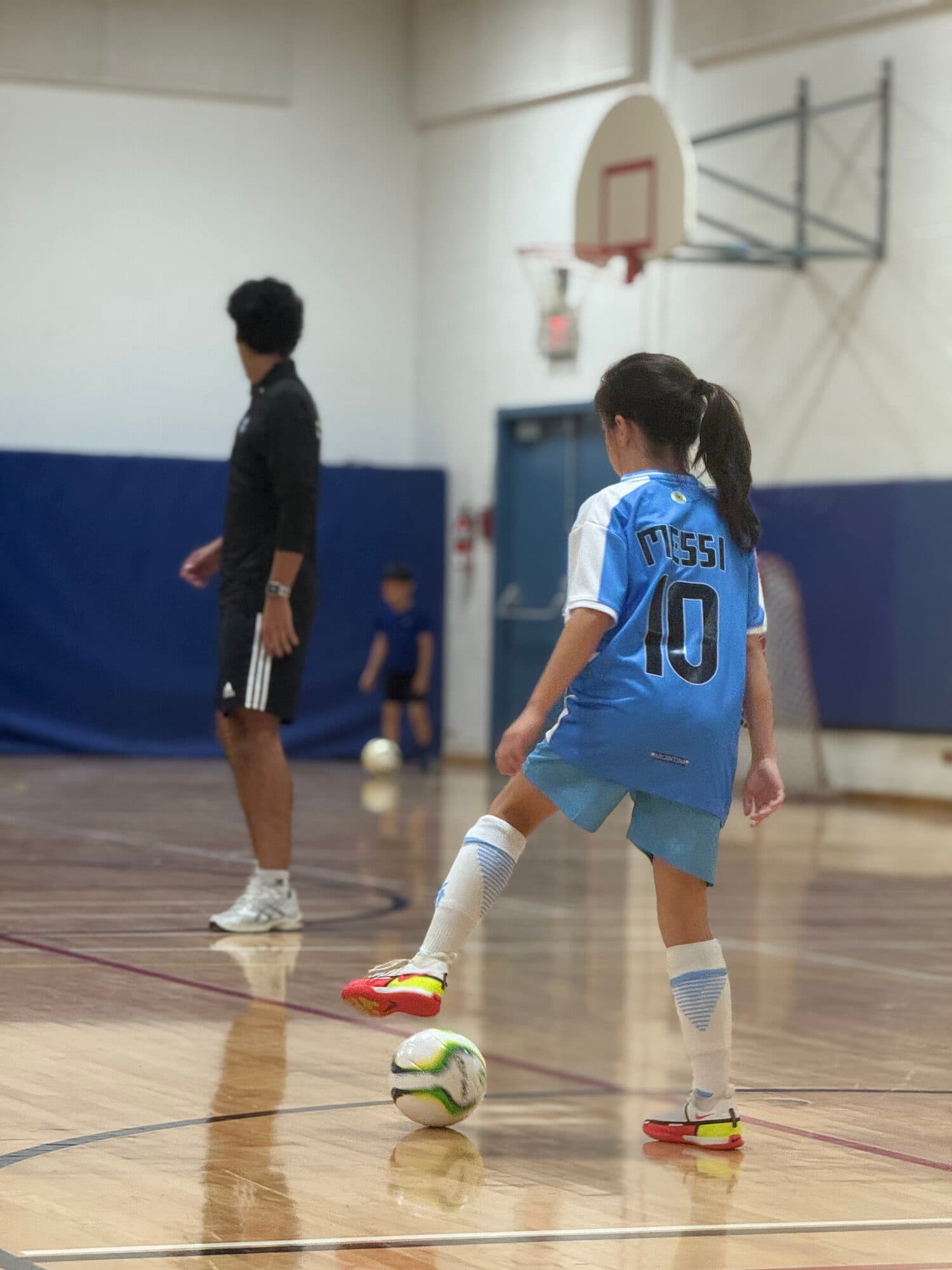 A young CFC player dribbles the ball in tight space during a training session, coach watching in the background
